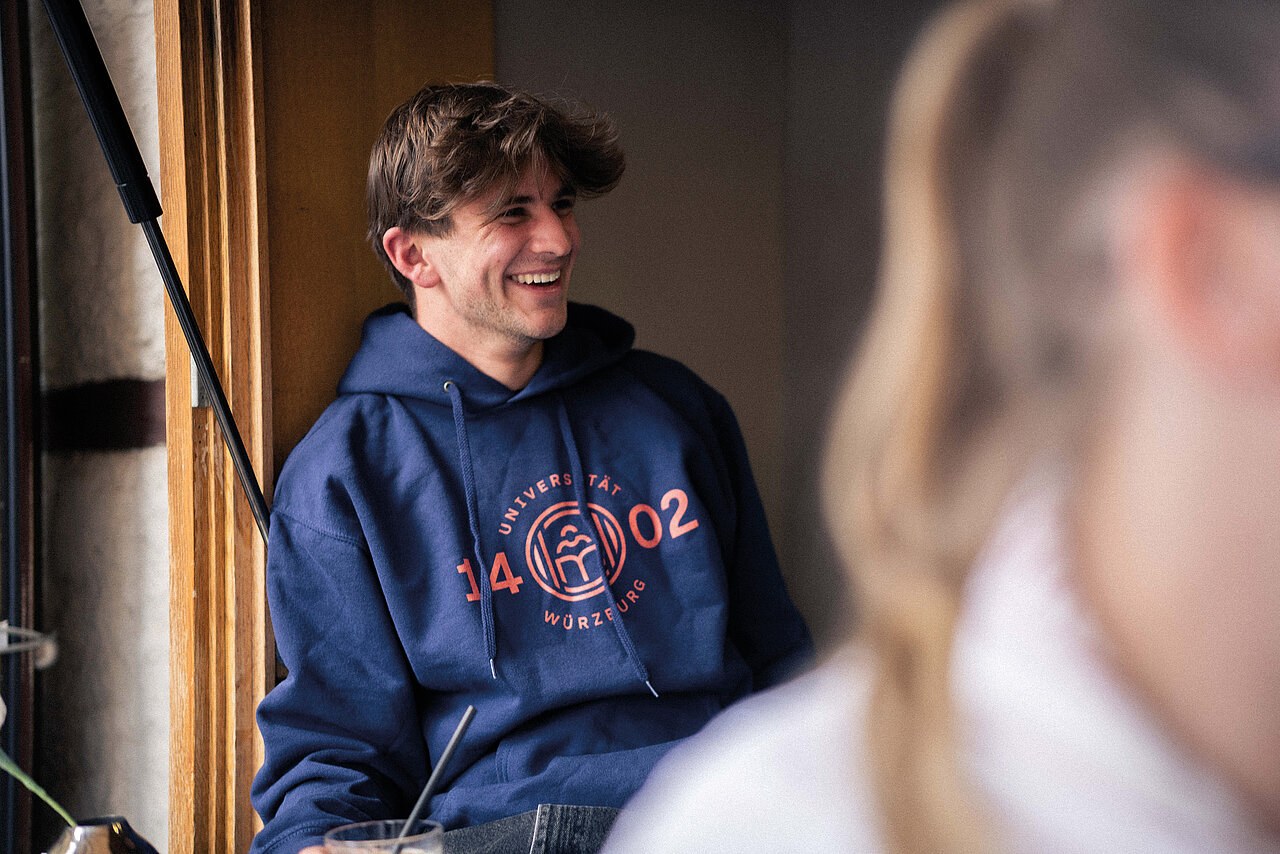Lachender Mann in einem blauen Hoodie mit rotem Siegel und der Zahl 1402
