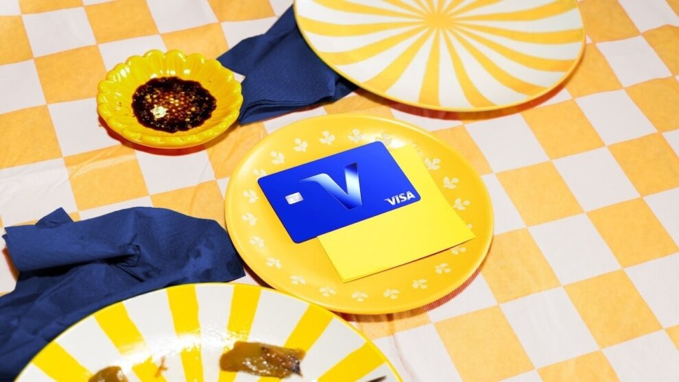 Blaue Visa-Karte mit großem V-Symbol auf gelbem Teller zwischen gelb-weißem Geschirr und blauen Servietten